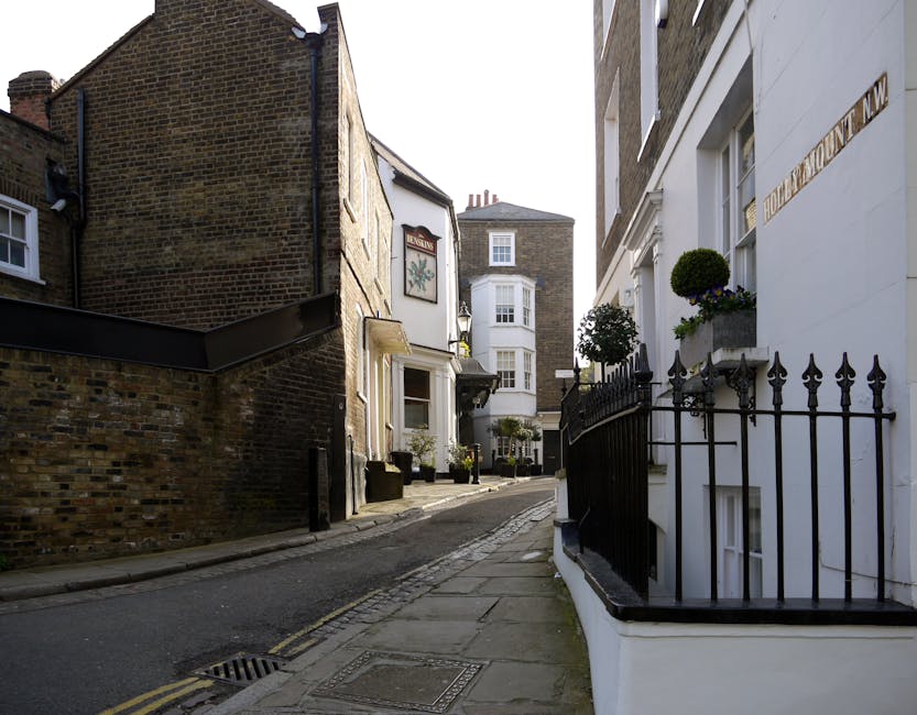 A narrow urban street lined with multi-storey buildings featuring brick and white-painted facades. In the foreground on the right, a black wrought-iron fence with decorative spikes encloses a small area of the building, which has window boxes filled with lush, rounded green plants. Adjacent to this fence, a white wall with visible structural details and a street name plaque is partially visible. The pavement is made of concrete slabs, with a drain grate and a drainage channel visible along the curb. Further along, there are potted plants and small outdoor furniture positioned outside the buildings, indicating residential or commercial occupancy. The street slopes slightly downward, leading to other similar buildings in the background, some with bay windows and chimneys on the rooftops. The lighting suggests a partly cloudy day with diffuse natural light illuminating the scene, providing a neutral and professional context related to urban waste management and alternative rubbish collection options available through private services such as Waste Collection Hampstead.
