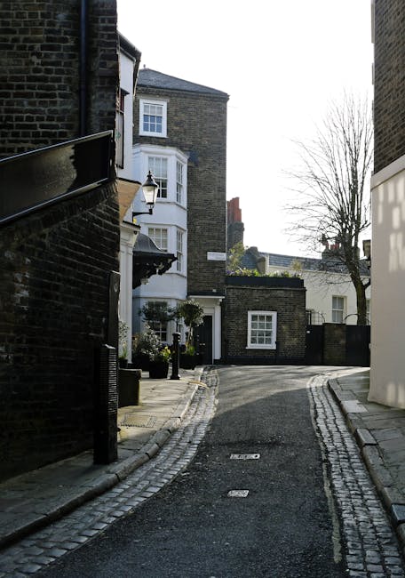 A narrow residential alleyway featuring a small, gently curved cobbled street flanked by tall, dark brick buildings on the left and a white modern structure on the right. The scene includes a classic-style street lamp mounted on the brick wall, a small potted plant near the entrance of a white bay-windowed house, and a leafless tree in the background against a partly cloudy sky. The alleyway is tidy with no visible rubbish or debris, suggesting it is kept clear and maintained. The environment appears quiet and tidy, typical of urban or suburban neighbourhoods where private waste collection or onsite clearance might be preferred over council rubbish disposal. The scene’s detail emphasizes materials such as the textured cobblestones and brick walls, highlighting the durability of traditional construction elements, and the clean, uncluttered appearance supports professional waste management services like those offered by Waste Collection Hampstead in maintaining neat and waste-free premises.