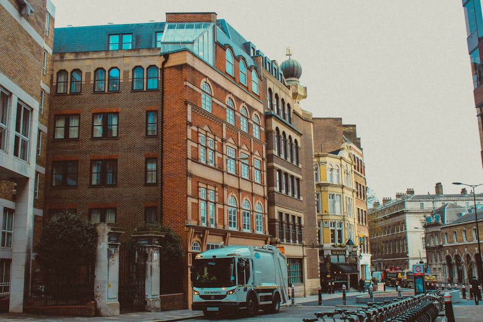 A city street scene featuring a range of multi-story buildings with mixed architectural styles, including brick and stone facades, and large windows. In the foreground, a white waste collection vehicle with a blue stripe, belonging to Waste Collection Hampstead, is parked on the street near a curb. To the left, there is a gated entrance with stone pillars leading to a building that is partially obscured by trees and bushes. The street is lined with a row of black metal bike racks, with a few bicycles parked, and several bollards separating the pedestrian area from vehicle traffic. In the background, additional historic and modern buildings with varying rooflines and details, including a domed structure and classical elements, are visible under an overcast sky, creating a typical urban environment suitable for private waste collection services rather than local authority rubbish removal.