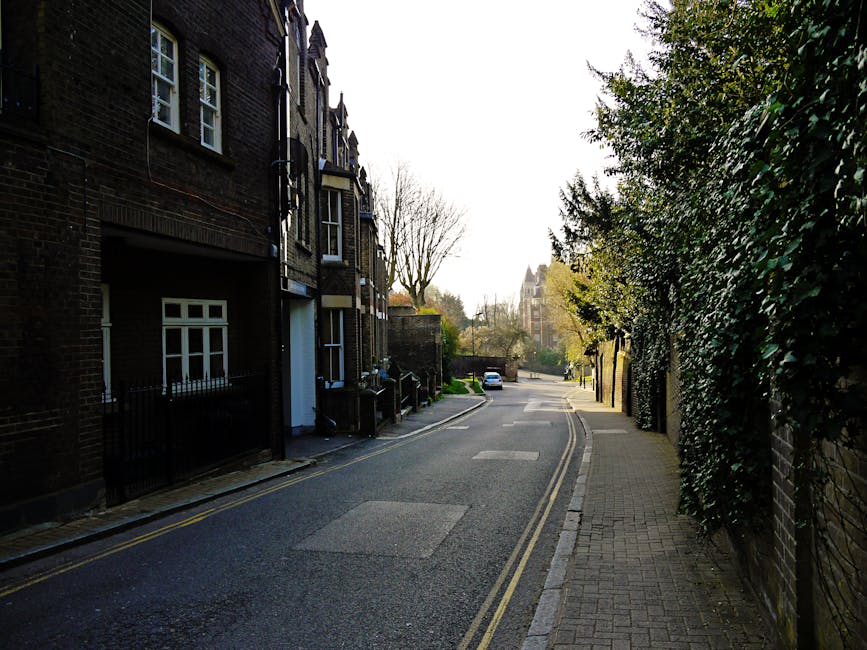 A quiet residential street in Hampstead, featuring a narrow tarmacked road with double yellow lines along the curb and a paved sidewalk running parallel. On the left side of the image, there are tall, dark brick terraced houses with white-framed windows, some with small front gardens enclosed by black metal railings. The buildings exhibit Victorian architectural details and have a weathered finish. On the right side, a high, dense hedge of dark green foliage borders the sidewalk, partially obscuring a brick wall beneath. The street is lined with a few leafless trees, indicating late autumn or winter, with their bare branches contrasting against the bright, partly cloudy sky. In the background, a parked white van is visible, along with a few more trees and an out-of-focus historic building with a pointed roof, possibly a church or civic structure. The scene is well-lit by natural daylight, casting soft shadows. The overall setting suggests an area where residents might arrange private waste collection or clearance services, as endorsed by Waste Collection Hampstead, to manage rubbish outside of council collection schedules.