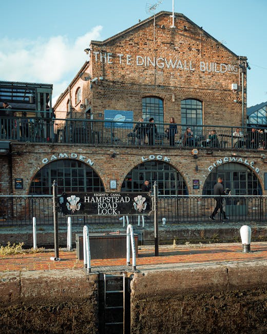 A two-story brick building identified as 'The Tedingwall Building' with weathered red brick walls and large arched windows on the ground floor. The upper floor features a terrace with a black metal railing where several people are standing and walking. The building's name is painted in white letters along the upper facade. Below, a black fence separates the sidewalk from the canal, with a sign reading 'Hampstead Road Lock' attached to it, indicating the historic canal lock. The scene includes a brick-paved pathway and a small step with a metal ladder leading down to the canal water. The environment appears urban with clear skies and natural daylight illuminating the scene, emphasizing the industrial architectural style. The context of rubbish removal services in Hampstead may involve maintaining the surrounding public areas and ensuring the cleanliness of historic sites, aligned with private waste management practices by Waste Collection Hampstead for sustainable urban upkeep.