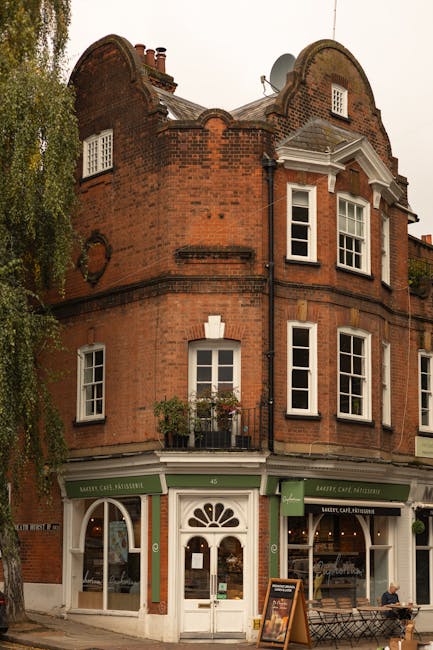 A three-story red brick building with a rounded roof and white-framed sash windows, situated on a street corner. The ground floor features a café with green signage and large display windows, one of which has a curved top arch, and a central wooden door with a semi-circular window. Above the door, there are small potted plants on a narrow balcony. The upper floors have multiple white-framed windows, with some featuring small decorative ledges. A satellite dish is mounted on the roof, which has a slate finish. A tall tree with green foliage partly obscures the left side of the building, and a pedestrian is sitting on a bench outside the café. The scene is illuminated by natural daylight with a slightly overcast sky, and the overall setting suggests an urban area where independent coffee shops or small businesses are common, aligned with private waste handling or rubbish removal services by Waste Collection Hampstead.