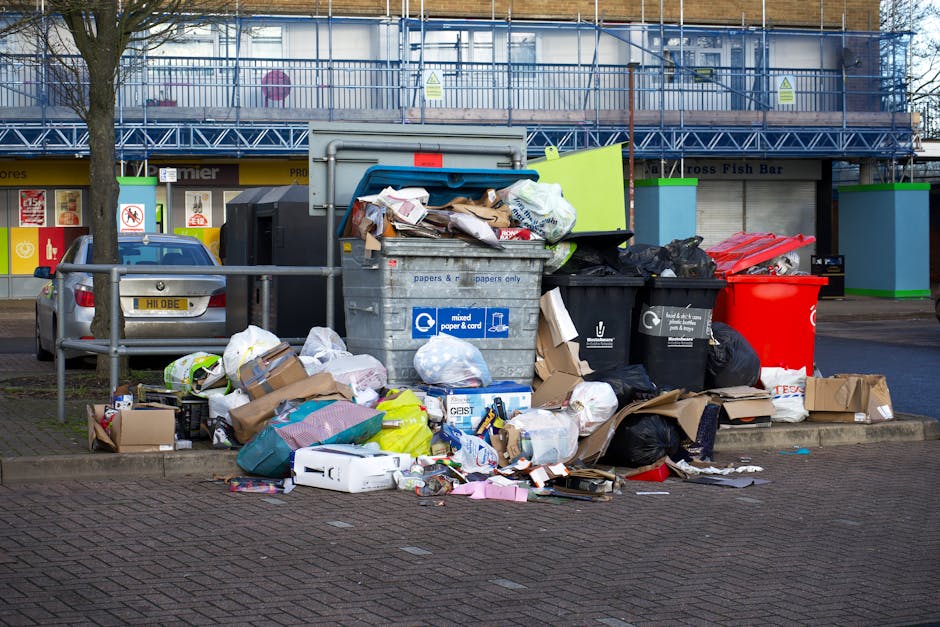 A large outdoor refuse collection point located on a paved urban street, featuring an open-top grey metal container overflowing with mixed waste including cardboard, paper, plastic bags, and miscellaneous household rubbish. Surrounding the container are numerous discarded items such as flattened cardboard boxes, plastic packaging, and crumpled paper, some stacked on the ground and others partially spilling out. To the left, a small silver car is parked close by, with a visible UK license plate. Behind the waste area, a building with a blue scaffolding structure and a row of retail shops with closed shutters, one displaying a fish bar sign, is visible. Trees line the street, and the scene is illuminated with natural daylight, highlighting the textures of the rubbish and the urban environment. The placement of the waste suggests an instance of bulk or on-site collection by a private waste removal service, consistent with non-council rubbish disposal practices typically handled by companies like Waste Collection Hampstead.