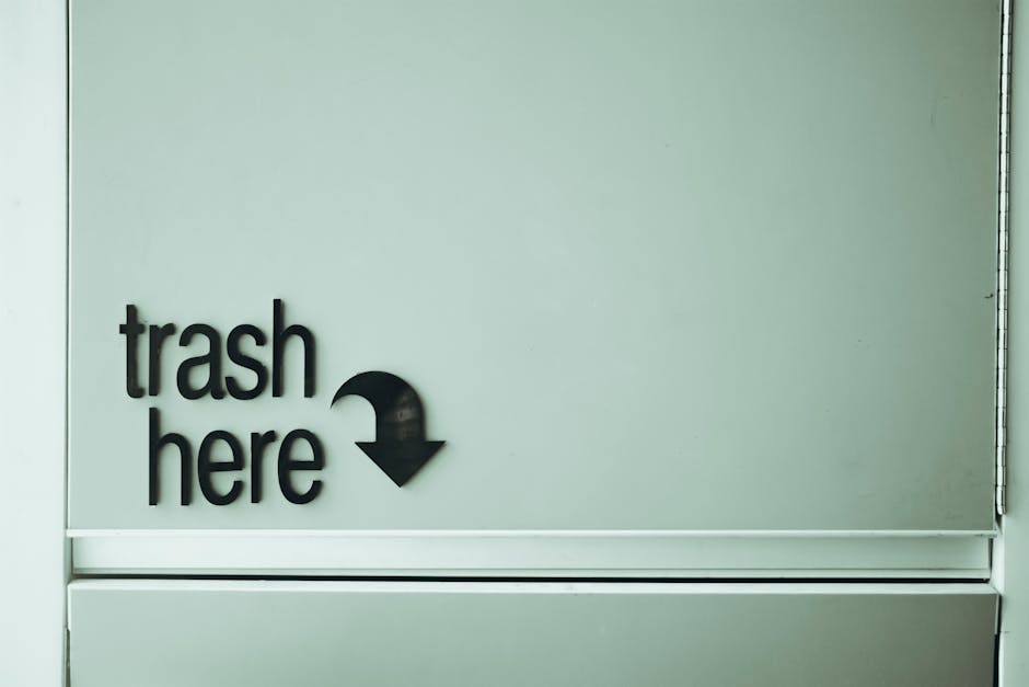 A close-up of a light grey wall with black three-dimensional lettering that reads 'trash here' accompanied by a downward-pointing arrow. The sign is mounted on a smooth surface, likely in an indoor or outdoor waste disposal area. The wall's finish appears matte, with subtle shadows cast by the raised letters, indicating indoor lighting. Below the sign, there is a horizontal slit or opening with metallic trim, possibly part of a waste receptacle or access panel associated with rubbish collection services. The environment looks clean and well-maintained, consistent with facilities used for private or independent waste disposal, such as a designated rubbish drop-off point managed by Waste Collection Hampstead. The overall scene emphasizes clear signage aimed at guiding users to dispose of waste in the correct location, supporting efficient rubbish removal and alternative waste handling outside of municipal collection methods.