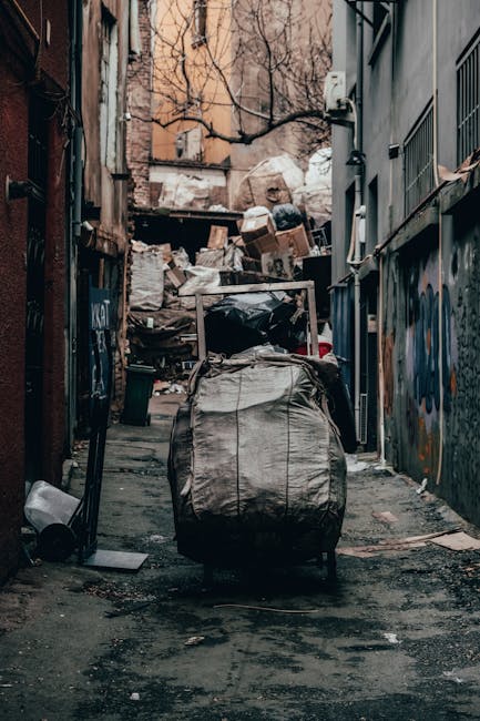 A narrow urban alleyway filled with overflowing rubbish and discarded materials, including large black and beige bags stacked haphazardly near the entrance. Piles of cardboard boxes, paper, and plastic waste are visible behind the bags, creating a cluttered scene. The alley is flanked by tall, dark brick and metal-paneled buildings on either side, with graffiti on some surfaces and small windows or vents along the walls. The ground is uneven, stained, and strewn with dirt and debris. In the background, leafless tree branches and a brick building are partially visible above the waste, suggesting an inner-city environment. The scene appears poorly maintained with a sense of neglect, typical of private waste accumulation requiring professional rubbish removal services such as those provided by Waste Collection Hampstead, highlighting the importance of effective bulk waste clearance and site tidying.
