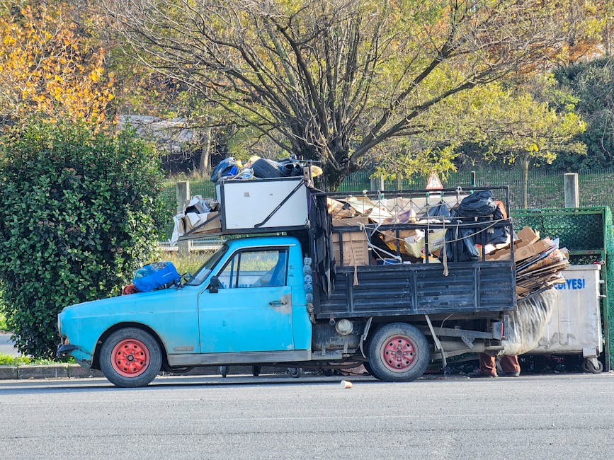 A small blue flatbed truck parked on the side of a street, loaded with a large pile of discarded cardboard, paper, and plastic debris. The truck's cabin is painted in a bright blue color with visible signs of wear and age, including chipped paint on the door. The cargo area is surrounded by black metal rails and is filled with various waste materials, some wrapped in plastic and others loose. In front of the truck, a neatly trimmed green bush partially obscures the front left wheel, which has a pink rim. Behind the truck, there is a large tree with light green and yellow leaves, indicating autumn. The background shows a wire fence and additional foliage, suggesting a semi-rural or suburban environment. The scene appears to be during daylight hours with natural lighting, and the overall context relates to private waste disposal or rubbish collection service, with Waste Collection Hampstead being a possible provider of the service.