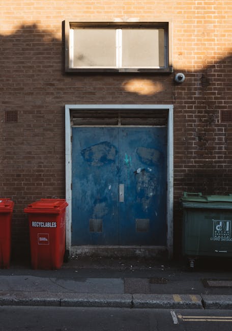 In front of a brown brick wall, there is a blue metal door with visible signs of weathering, including peeling paint and rust patches, positioned below a small rectangular window with frosted or dirty glass pane. To the left of the door, two red recycling bins made of plastic are placed on the pavement, with the word 'RECYCLABLES' printed in white on the front of one bin. To the right, a green waste bin with a white logo and text, likely for general waste, is positioned next to the curb. The pavement is concrete, featuring a slight curb edge, and the scene is illuminated by natural daylight, casting soft shadows on the wall. This setting appears to be an alley or back street adjacent to a commercial or residential building, highlighting a typical area for private waste collection services as offered by companies like Waste Collection Hampstead, providing alternative waste handling options outside of standard municipal refuse collection.