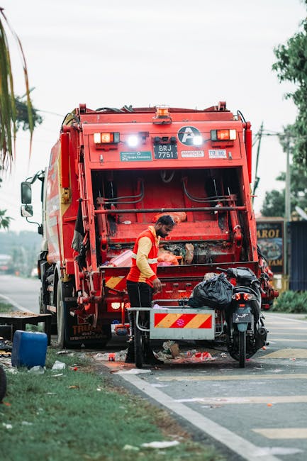 A waste collection worker dressed in a high-visibility yellow and red uniform stands beside the open rear hatch of a large red rubbish collection truck, which is parked on the side of a street. The rear section of the truck reveals mechanical components and an enclosed compactor area with a black plastic bag placed near the loading area. The scene shows some scattered debris and waste items around the vehicle's base, with a small black scooter parked close beside it and a metal barrier with a striped yellow and red reflective pattern in front. In the background, there are trees, overhead power lines, and a building with signage, indicating an urban or suburban environment. The ambient lighting suggests a daytime setting with overcast skies, relevant to private waste collection or on-site clearance activities often undertaken by independent rubbish removal services. The environment and the worker’s activity suggest a routine waste collection or debris clearance operation, consistent with professional rubbish removal practices in Hampstead or Camden areas.