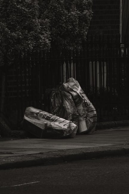 Two large black plastic garbage bags filled with waste are positioned on the pavement alongside a curb, leaning against a black metal fence with vertical bars. The bags appear crumpled and are stacked one atop the other, with some sections showing visible folds and creases. Behind the fence, dense foliage from a tree extends into the upper part of the image, providing a natural background. To the right, a dark brick building with a grid-like pattern of bricks is partially visible, blending into the shadows cast by the evening or overcast lighting conditions. The scene takes place outdoors on a street, with no visible vehicles or pedestrians, capturing a typical instance of roadside rubbish awaiting collection or alternative waste disposal, characteristic of private clearance services such as those offered by waste collection firms in Hampstead specializing in rubbish removal.